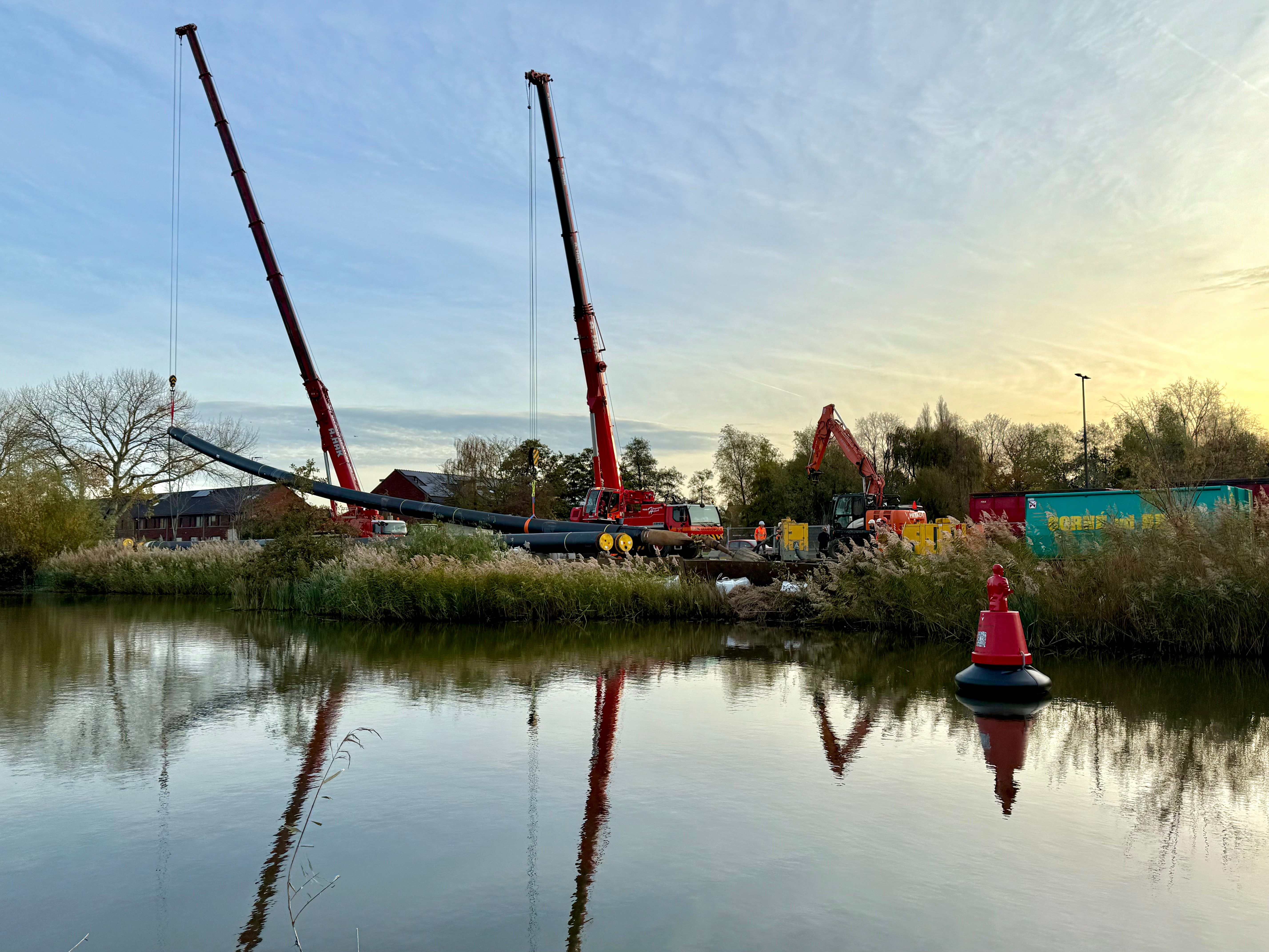 Twee kranen houden het leidingdeel in de lucht zodat deze onder de juiste hoek de grond in wordt getrokken.