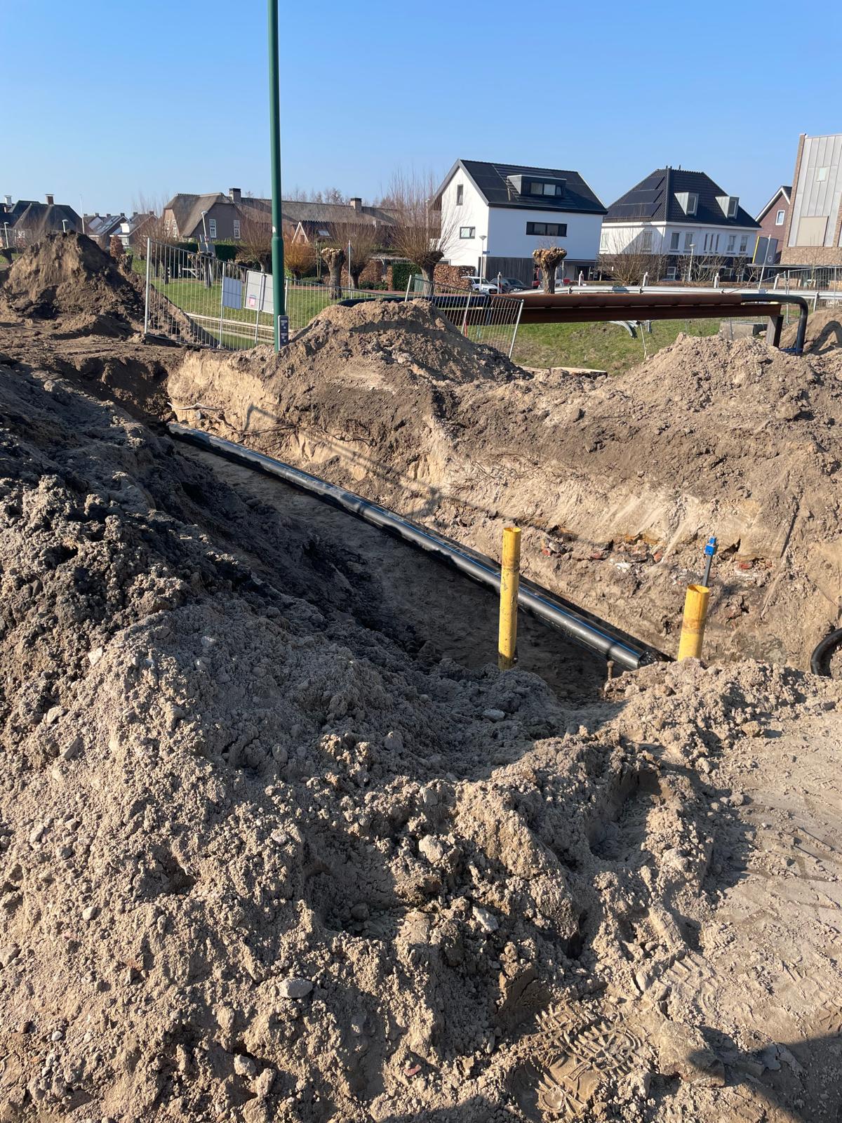 Gegraven sleuf op de Rijksstraatweg met een nieuw geplaatst leidingdeel, omringd door opgegraven zand en hekwerken.