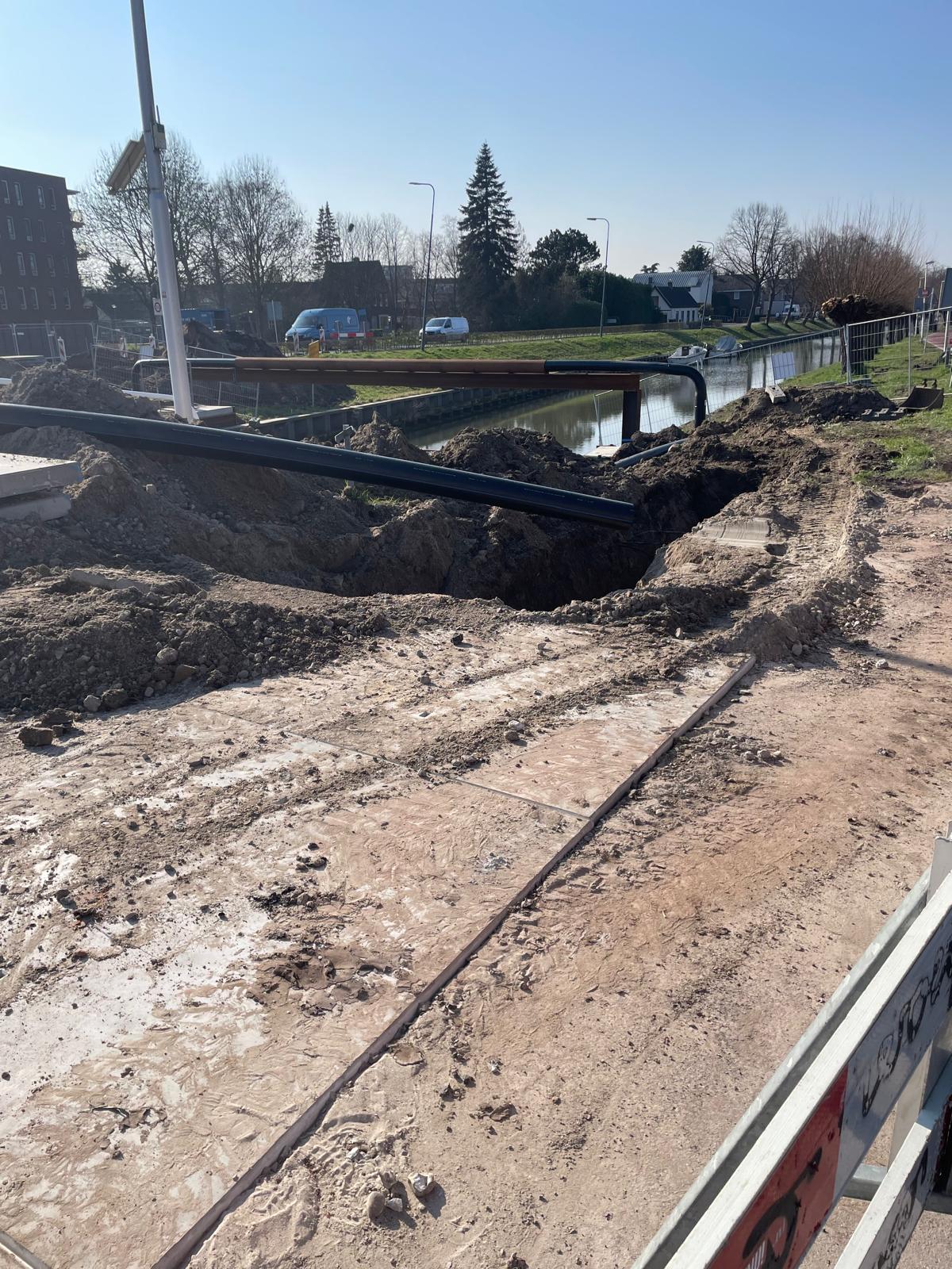 Grondwerk ter hoogte van de Zandweg waar een sleuf is gegraven voor het omleggen van de waterleiding, met een tijdelijk portaal boven de watergang.