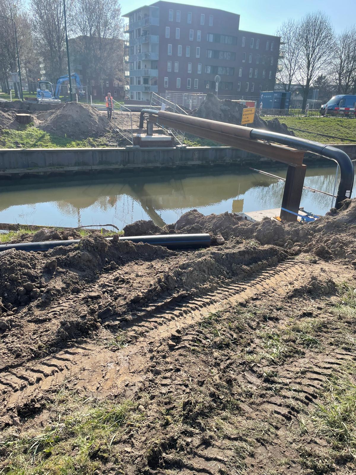 Werkzaamheden langs de watergang waar een tijdelijke leidingconstructie over het water ligt, met machines en bouwmateriaal op de achtergrond.