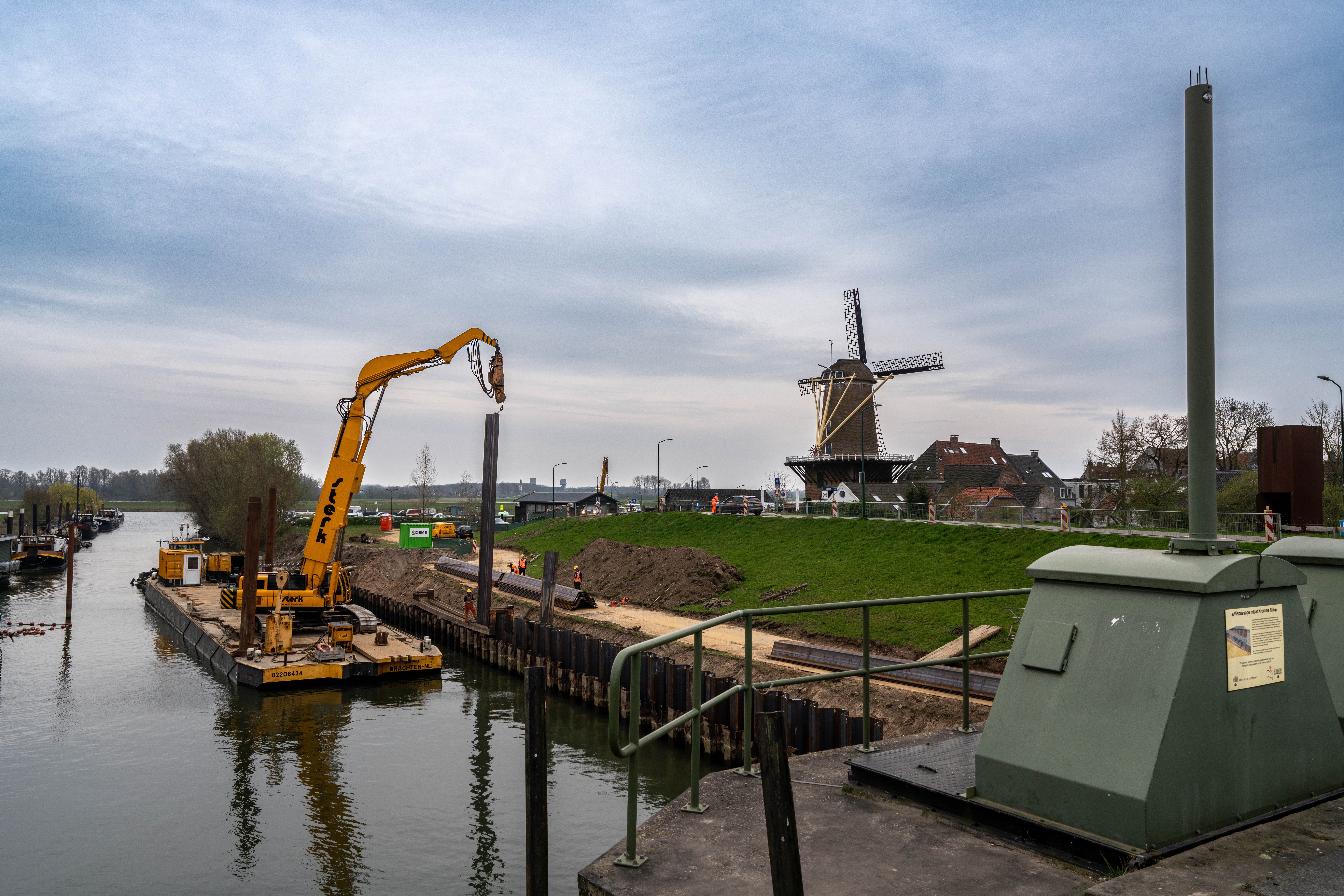 Aanzicht vanaf de sluis op de ponton met de kraan die damwanden plaatst, met de historische molen op de achtergrond.